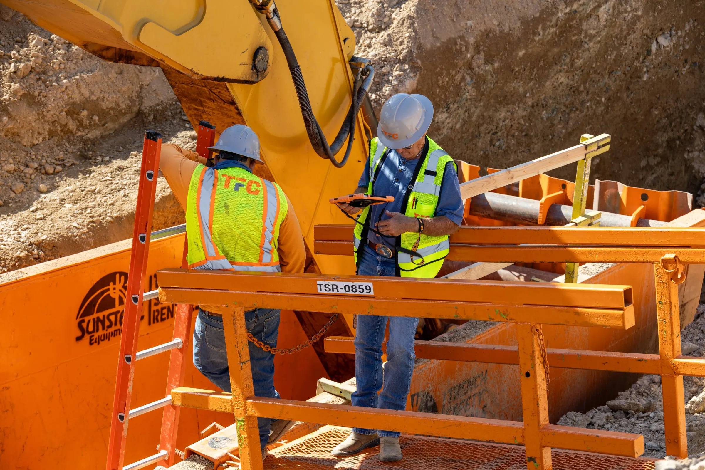 Two workers in hard hats and safety vests reviewing plans in a trench box